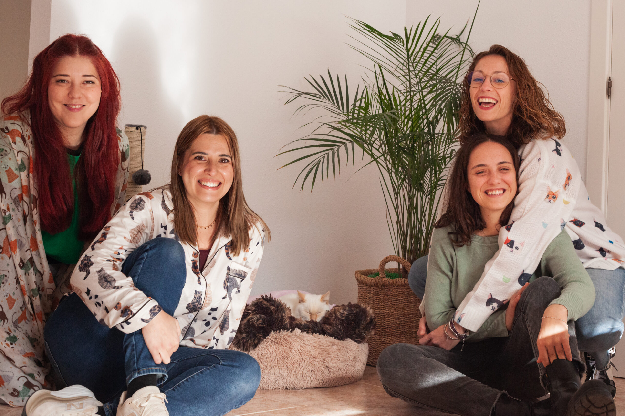 Foto QS Cuatro mujeres sonrientes sentadas en el suelo de una habitación luminosa con una planta al fondo. Van vestidas de manera informal, algunas con camisas de estampado animal. Un gato blanco con manchas naranjas duerme en una cama peluda entre ellas. "GEMMA LAHOZ fotografía" en la esquina inferior derecha.